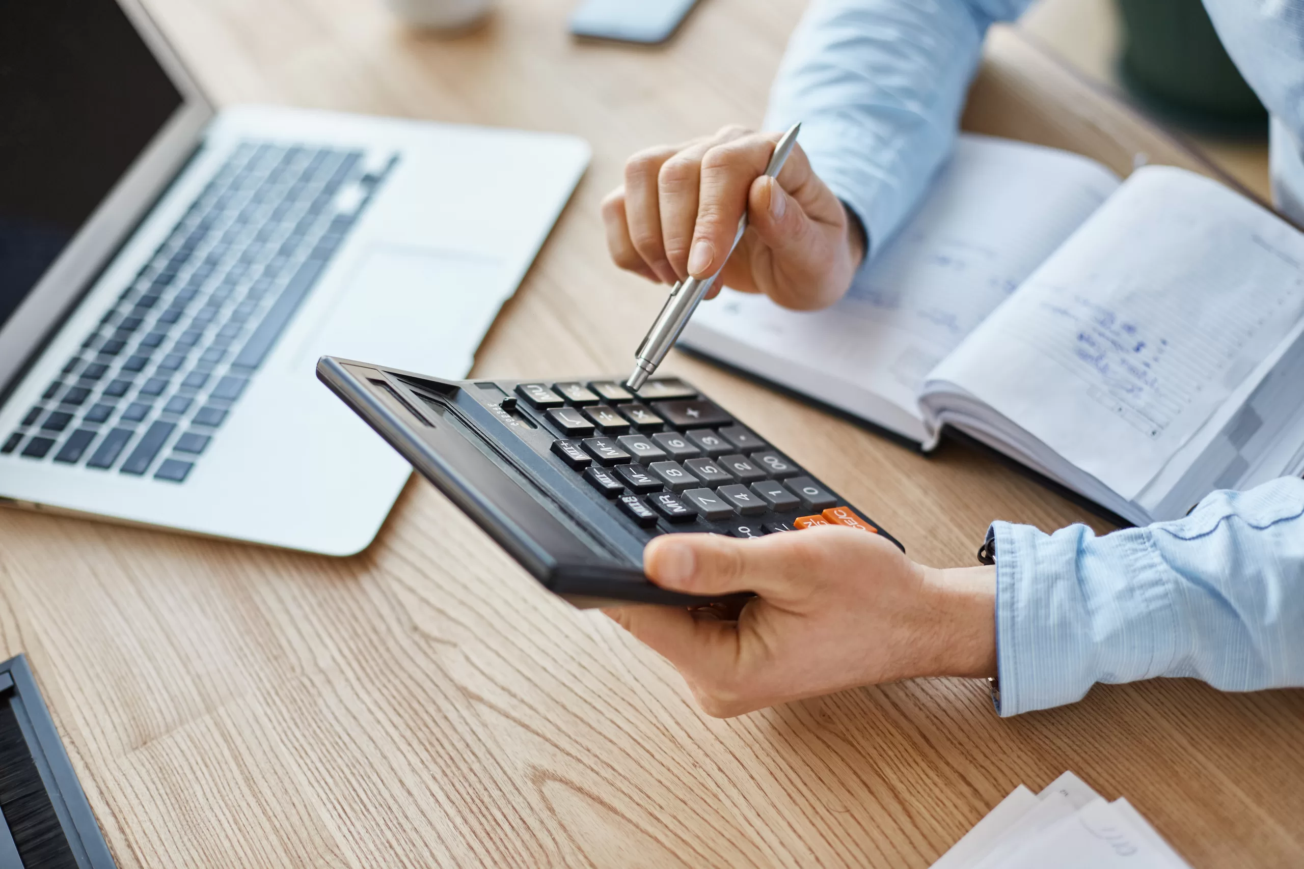 Cropped view of professional serious finance manager, holding calculator in hands, checking company month's profits, looking through details on laptop, writing down information for meeting with director.